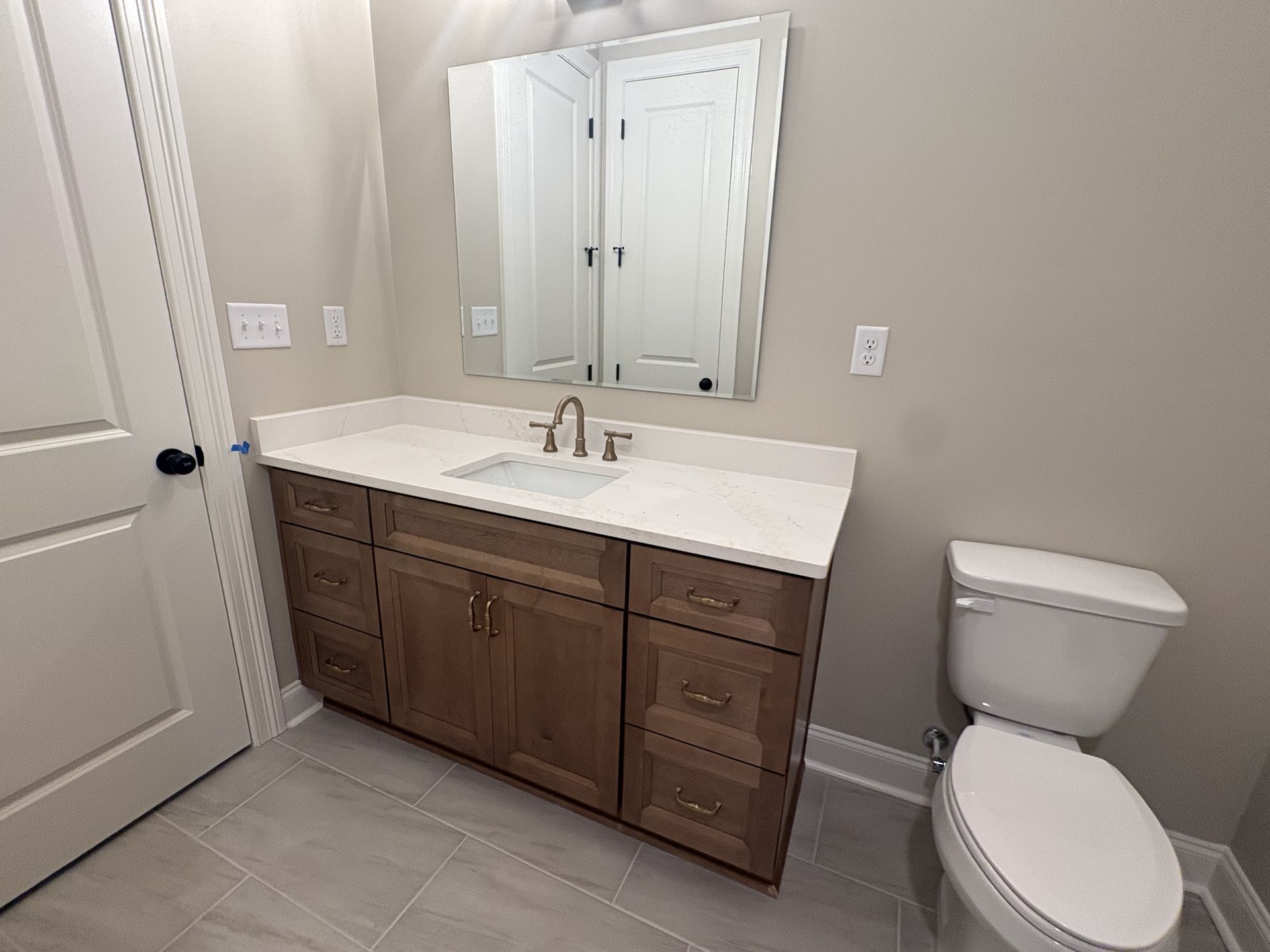 Bathroom with wooden vanity, white countertop, mirror, and toilet on beige tile floor.