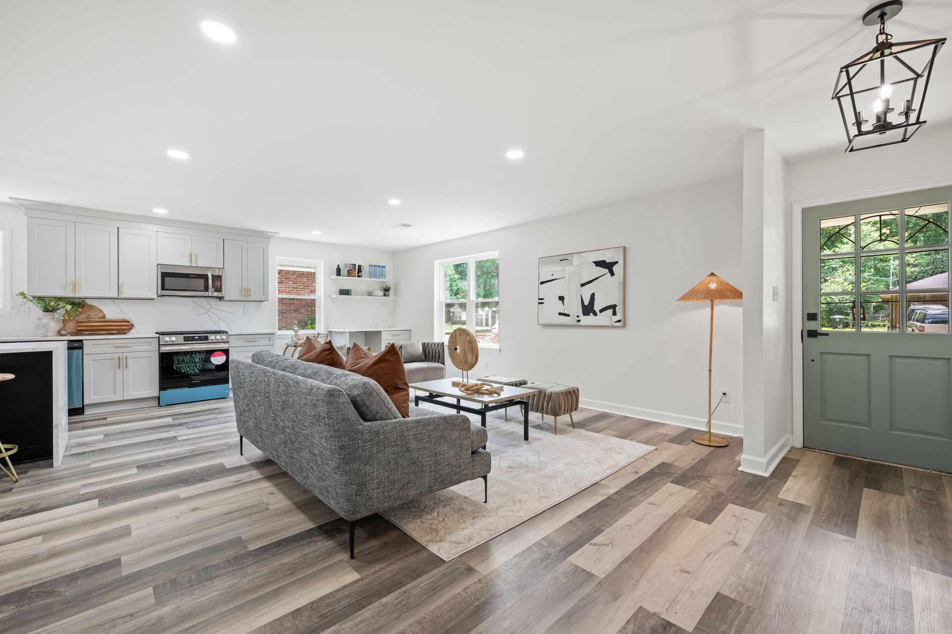 Open-concept living space with kitchen and living room. Gray sofa, light wood floors, and sage green door.