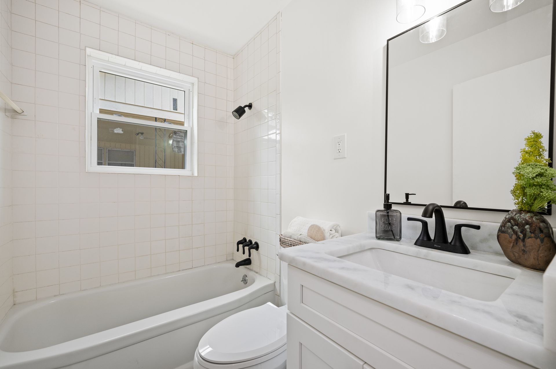 White bathroom with tub, toilet, vanity, and window. Black fixtures and marble countertop.
