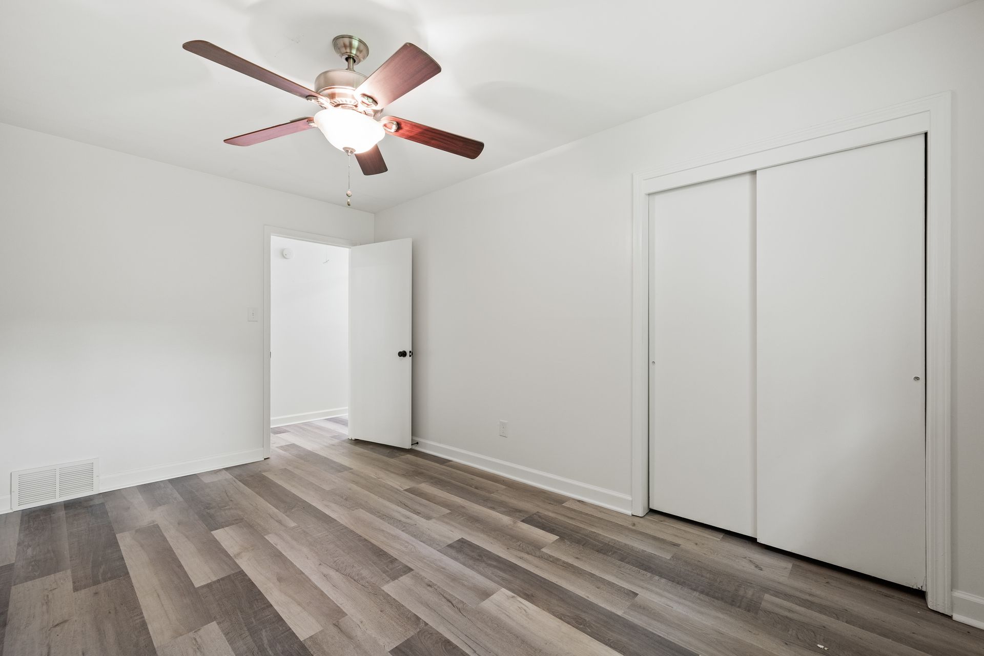 Empty bedroom with white walls, sliding closet doors, and wood-look flooring. A ceiling fan is installed.