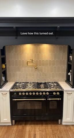 Black stove with gold accents, beige tile backsplash, cream cabinets, and wooden floors.