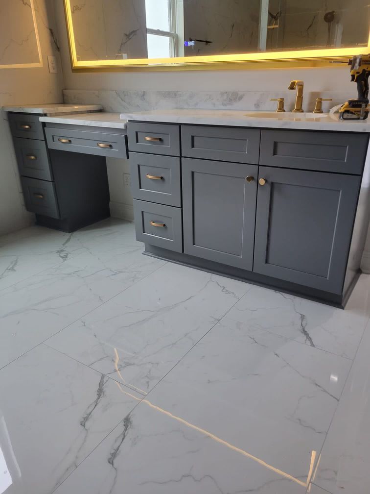 Gray bathroom vanity with white marble countertop and tile floor, gold fixtures.