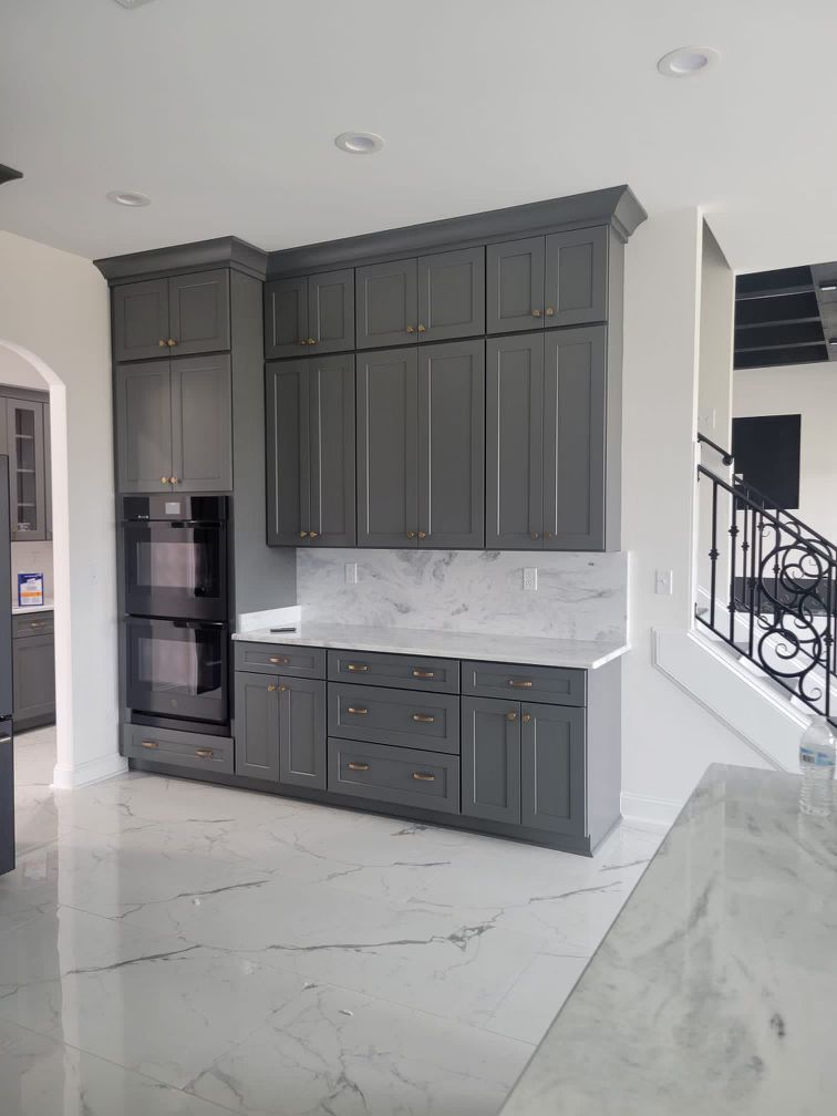Gray kitchen cabinets with double ovens and marble backsplash. White marble floors.