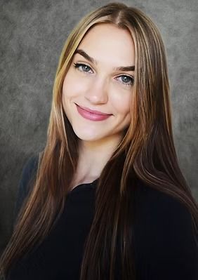 Woman with long brown hair smiling at the camera, wearing a black shirt, against a grey backdrop.