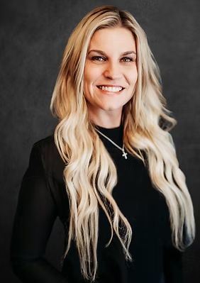 Blonde woman smiling, wearing black top, posing against dark background.