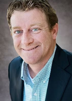 Man in suit jacket and button-down shirt smiles, looking at the camera.