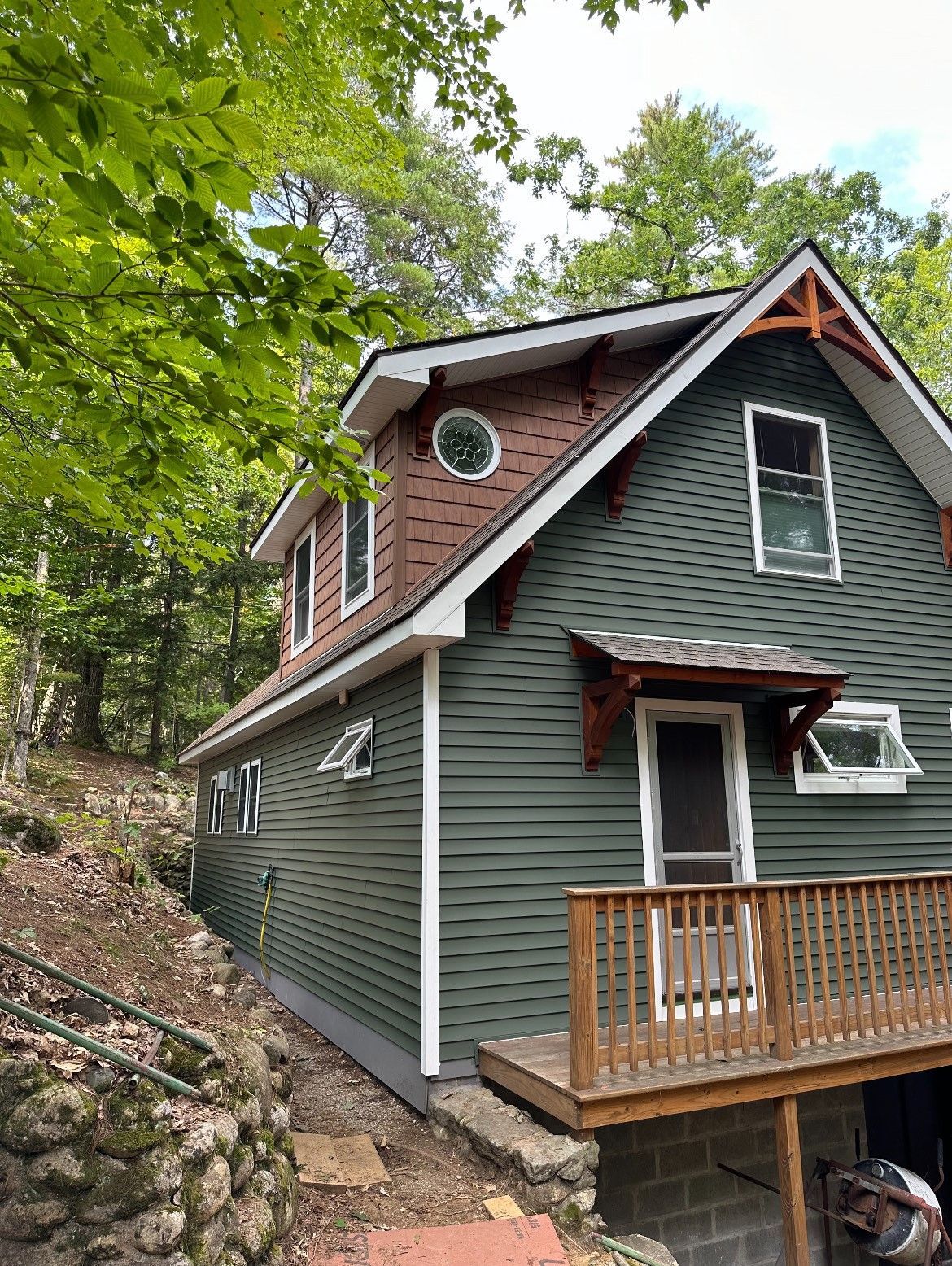A green house with a brick roof and a deck in the woods.