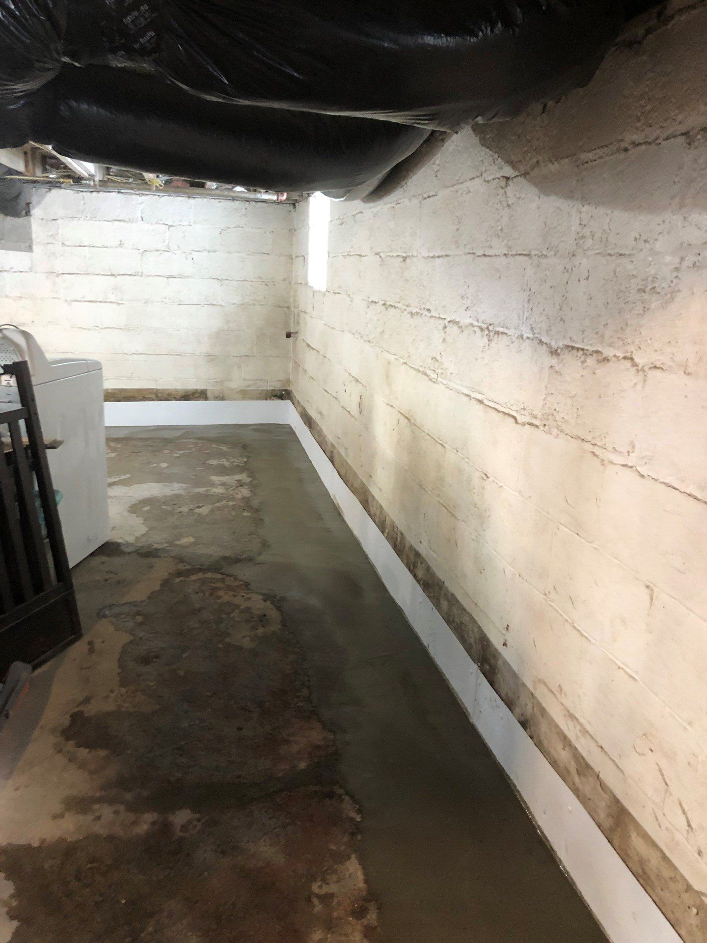 Narrow basement corridor with damp concrete floor, white block walls, and low black plastic sheeting overhead