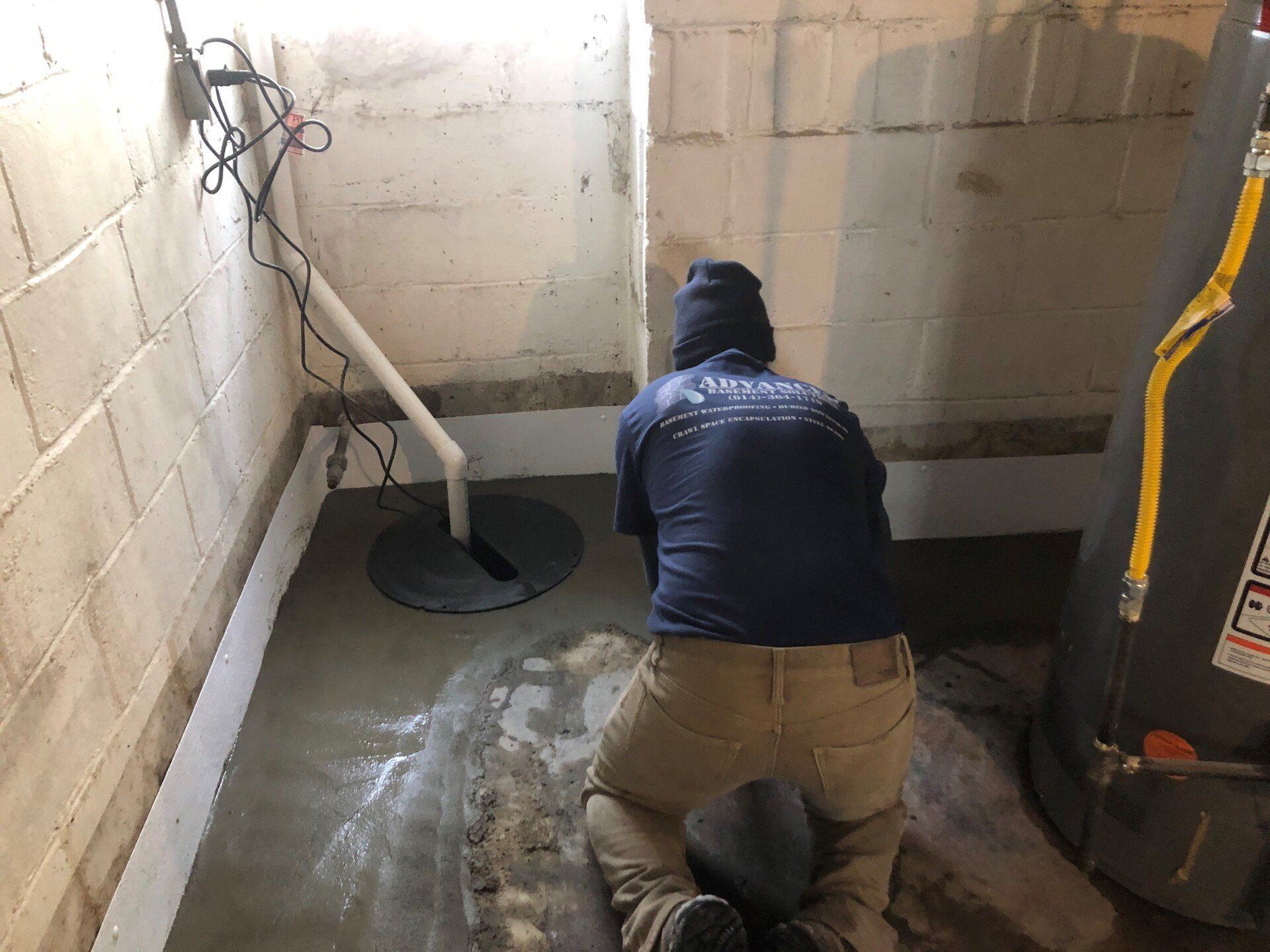 Worker kneeling in a basement near a water heater, applying sealant to the concrete floor.