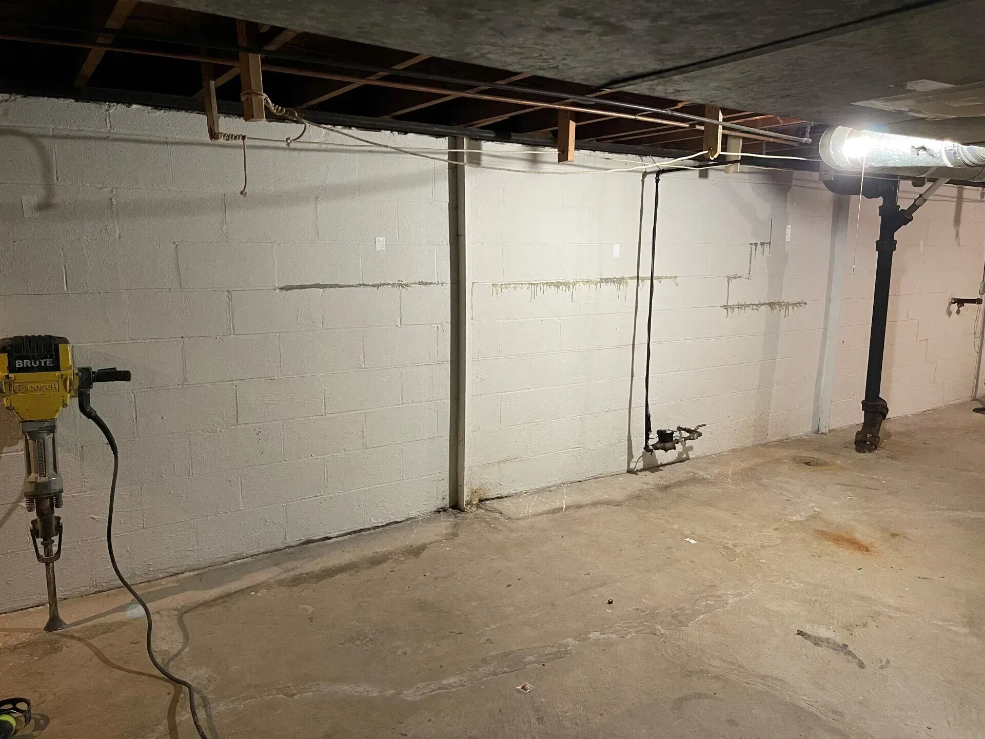 Basement with cinderblock wall, exposed pipes and wiring, and a small work light on the ceiling.