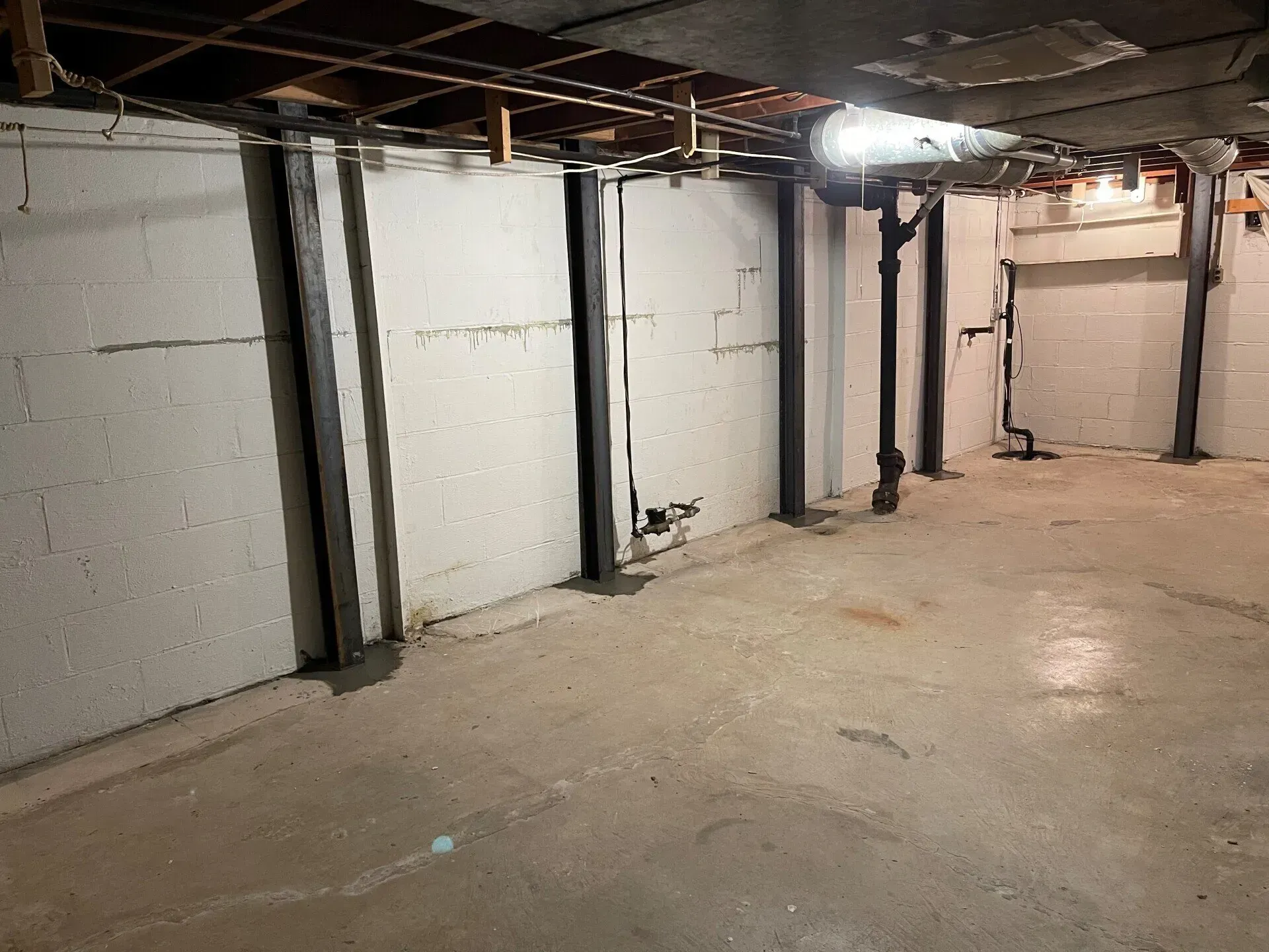 Empty unfinished basement with cinder block walls, exposed ceiling pipes, and concrete floor under one light