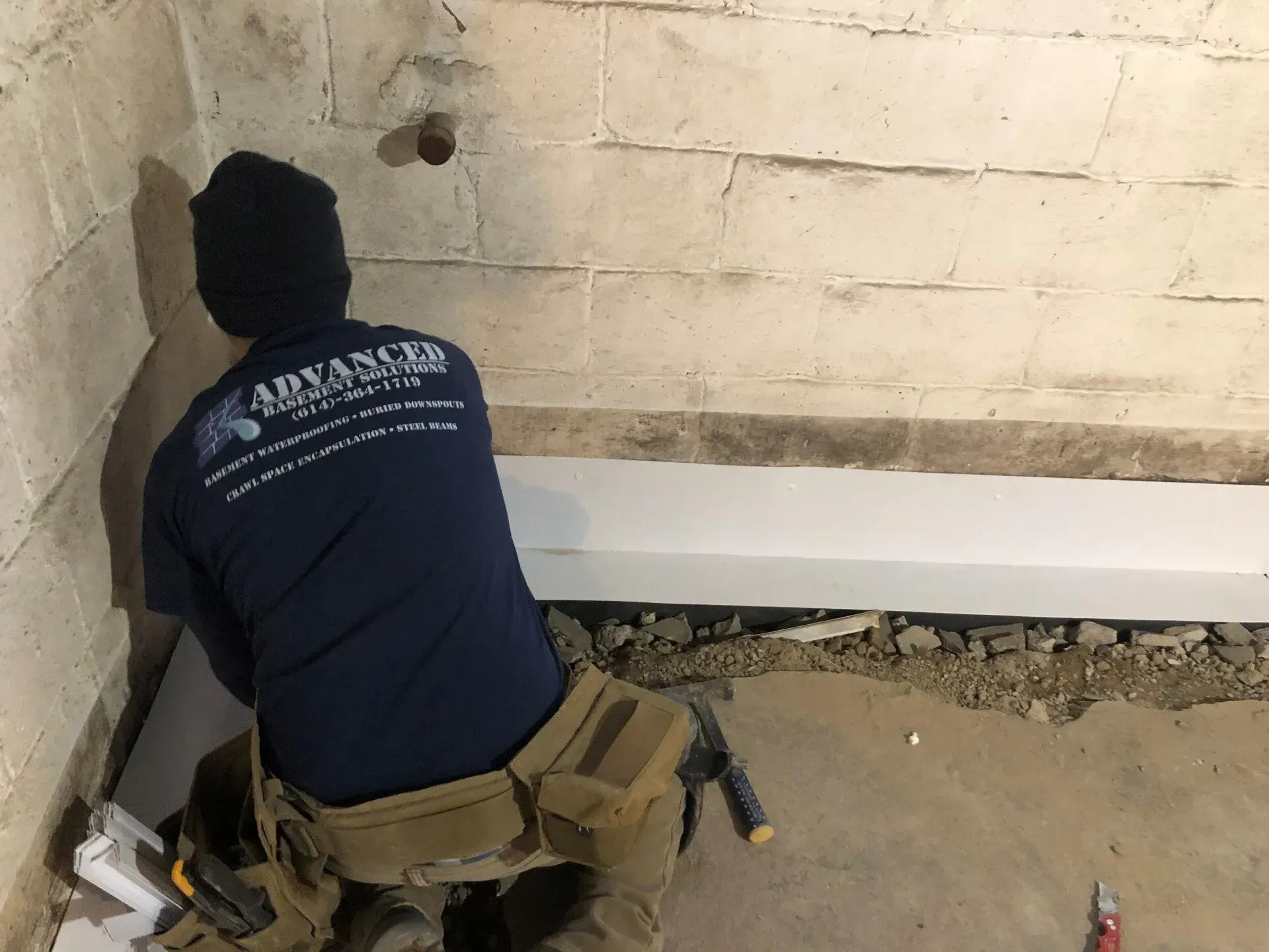 Worker crouching by a wall, using tools to inspect or repair a damaged floor edge.