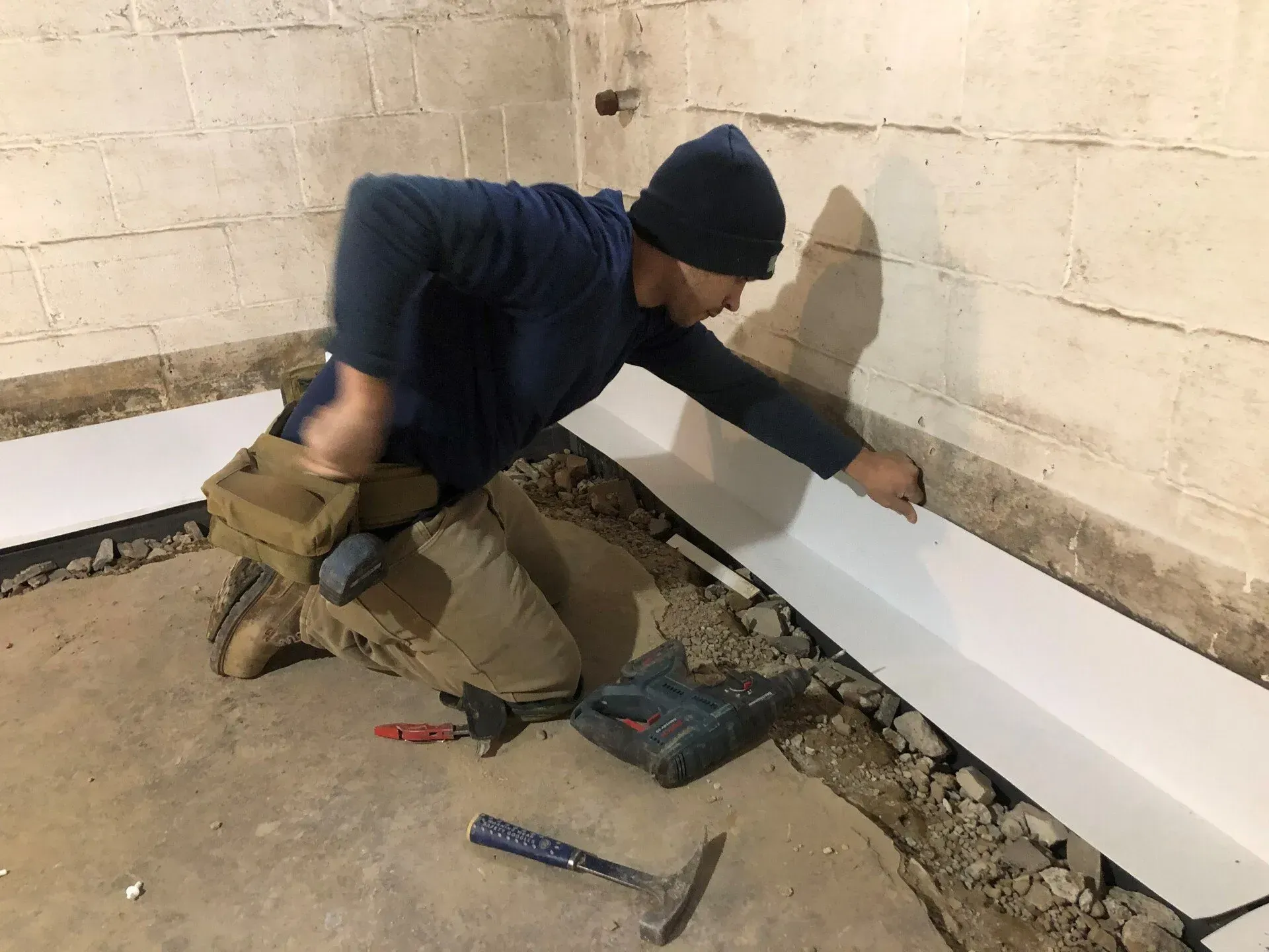 Worker installing white baseboard along a basement wall, kneeling with tools and debris nearby