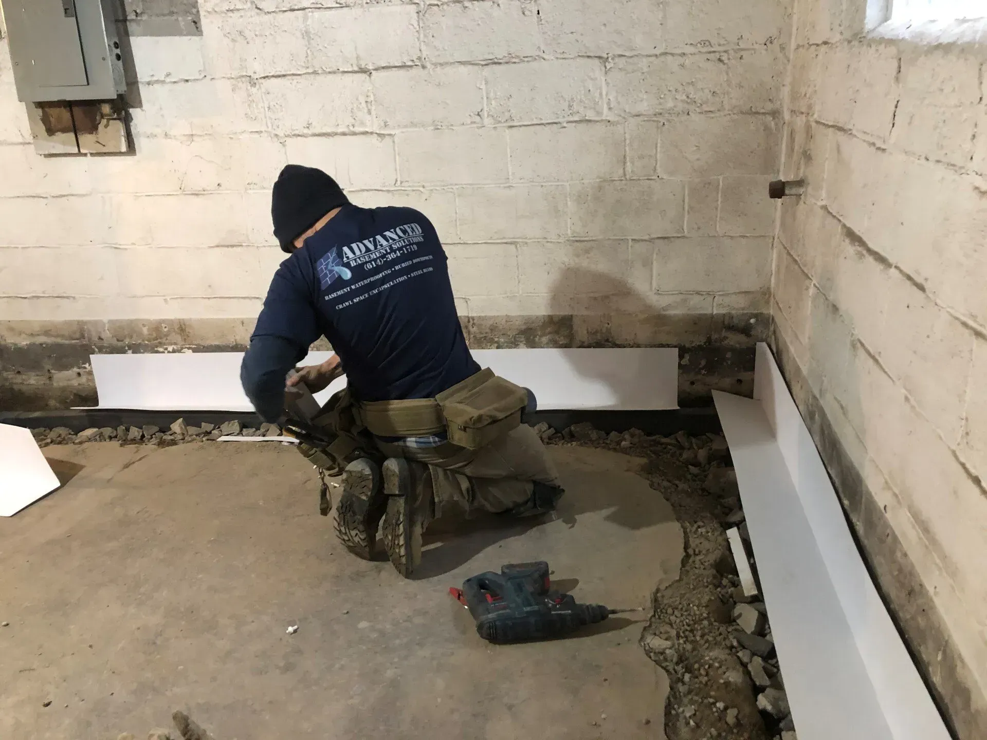 Worker kneeling in a basement, applying mortar along the wall floor joint with tools nearby.