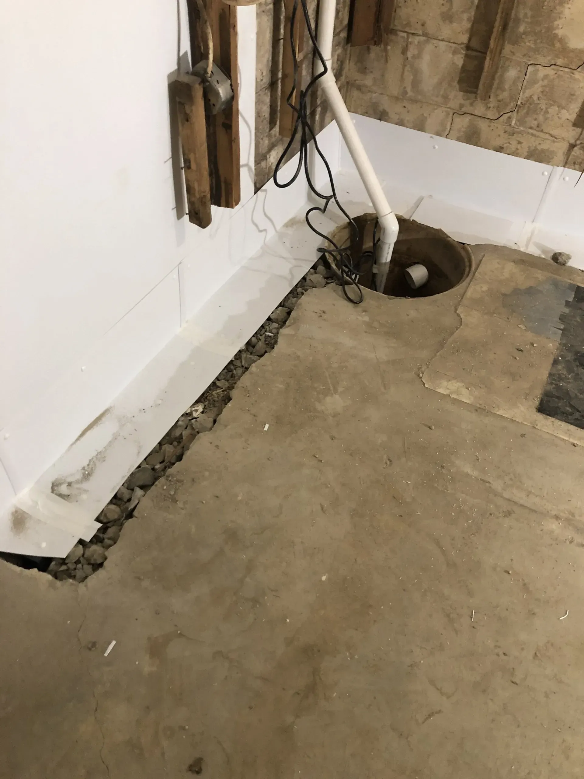 Basement corner with exposed wires, pipes, and a floor hole next to a white wall and concrete floor