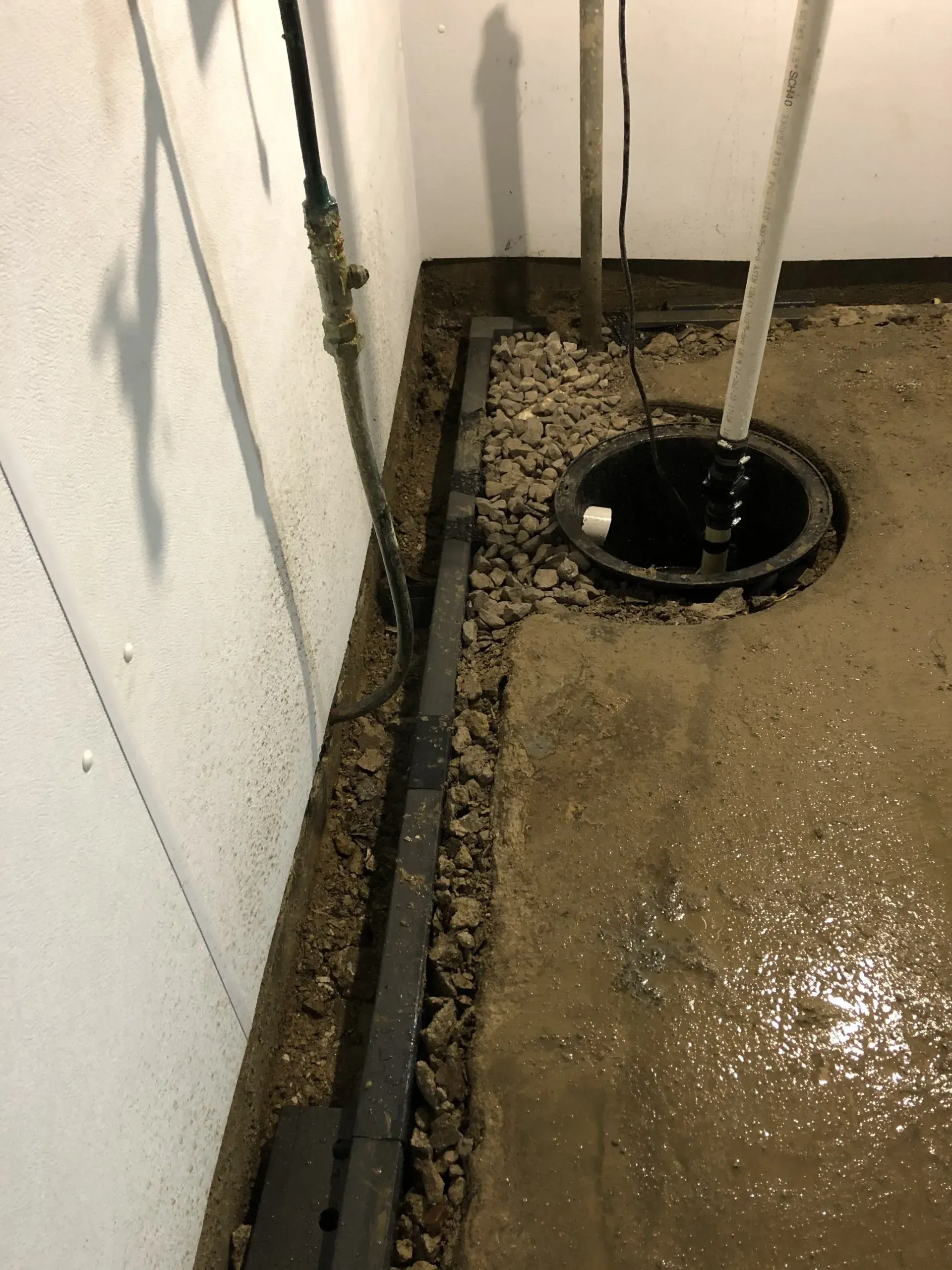 Utility trench beside a wall with gravel, pipes, and a round drain opening on a wet concrete floor