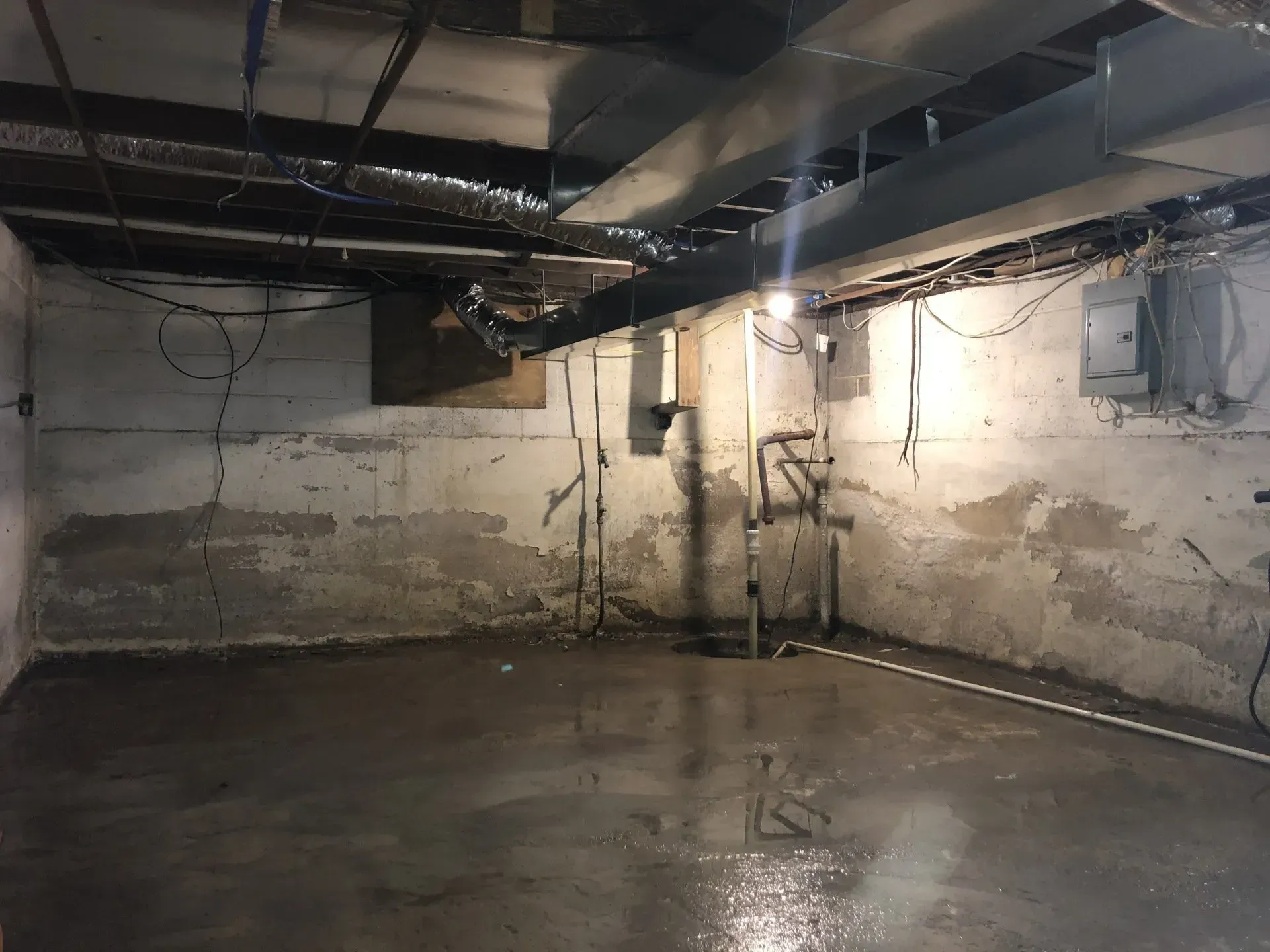 Empty unfinished basement with concrete walls, exposed ceiling pipes, and a wet floor.
