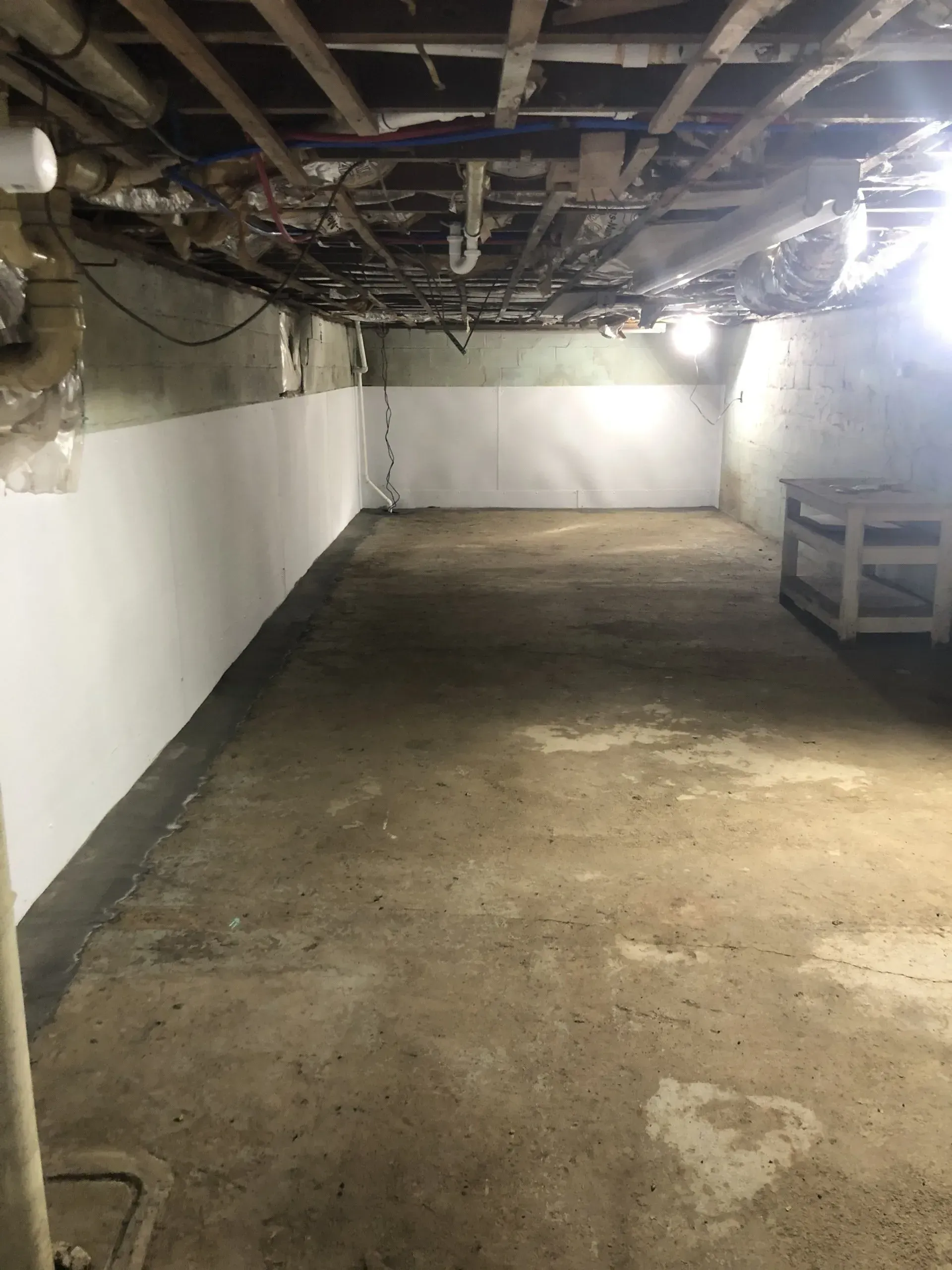 Dim basement crawlspace with white walls, exposed pipes, concrete floor, and a workbench on the right.
