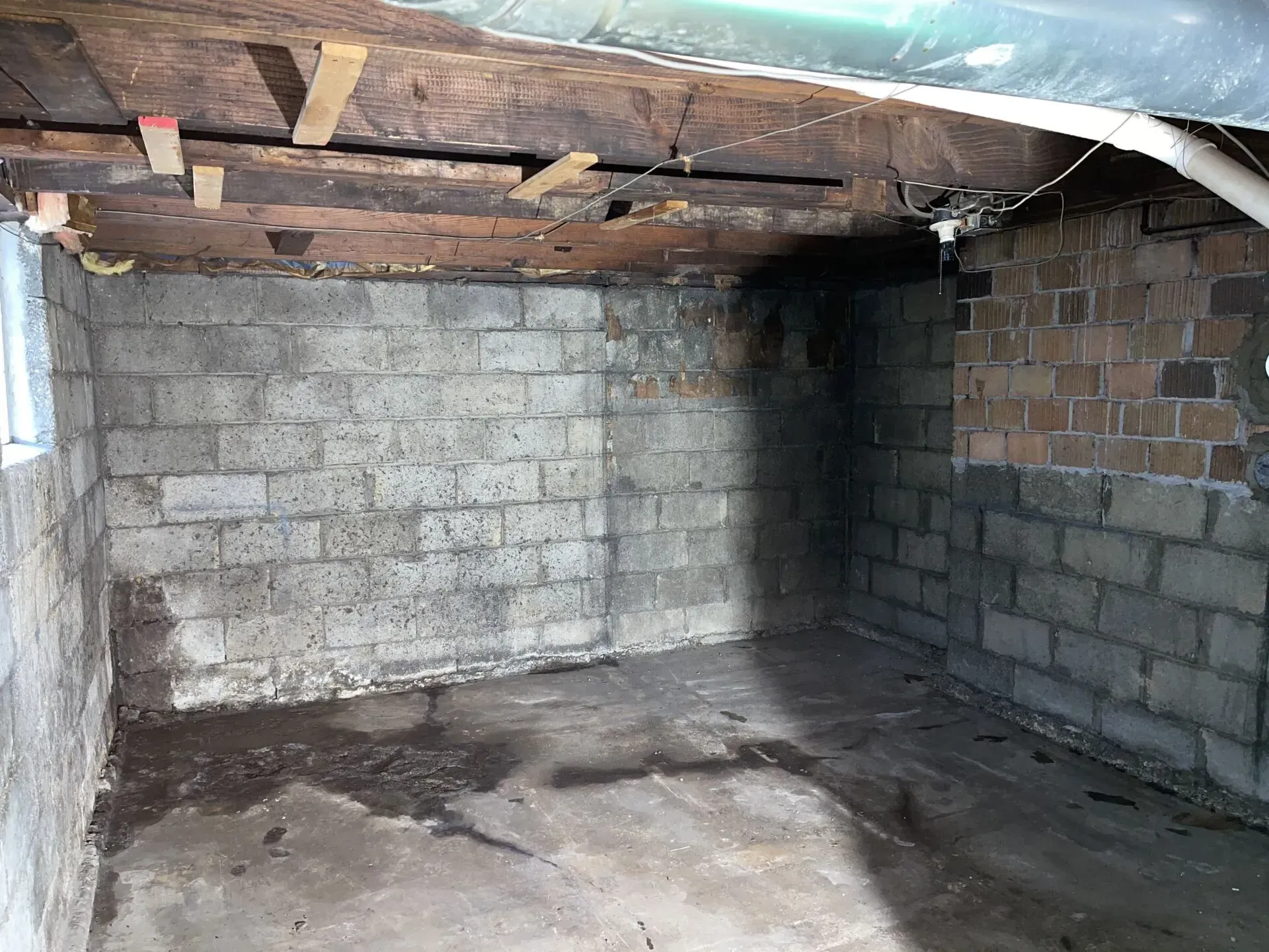 Empty basement with cinder block walls, exposed wooden ceiling beams, and a damp concrete floor