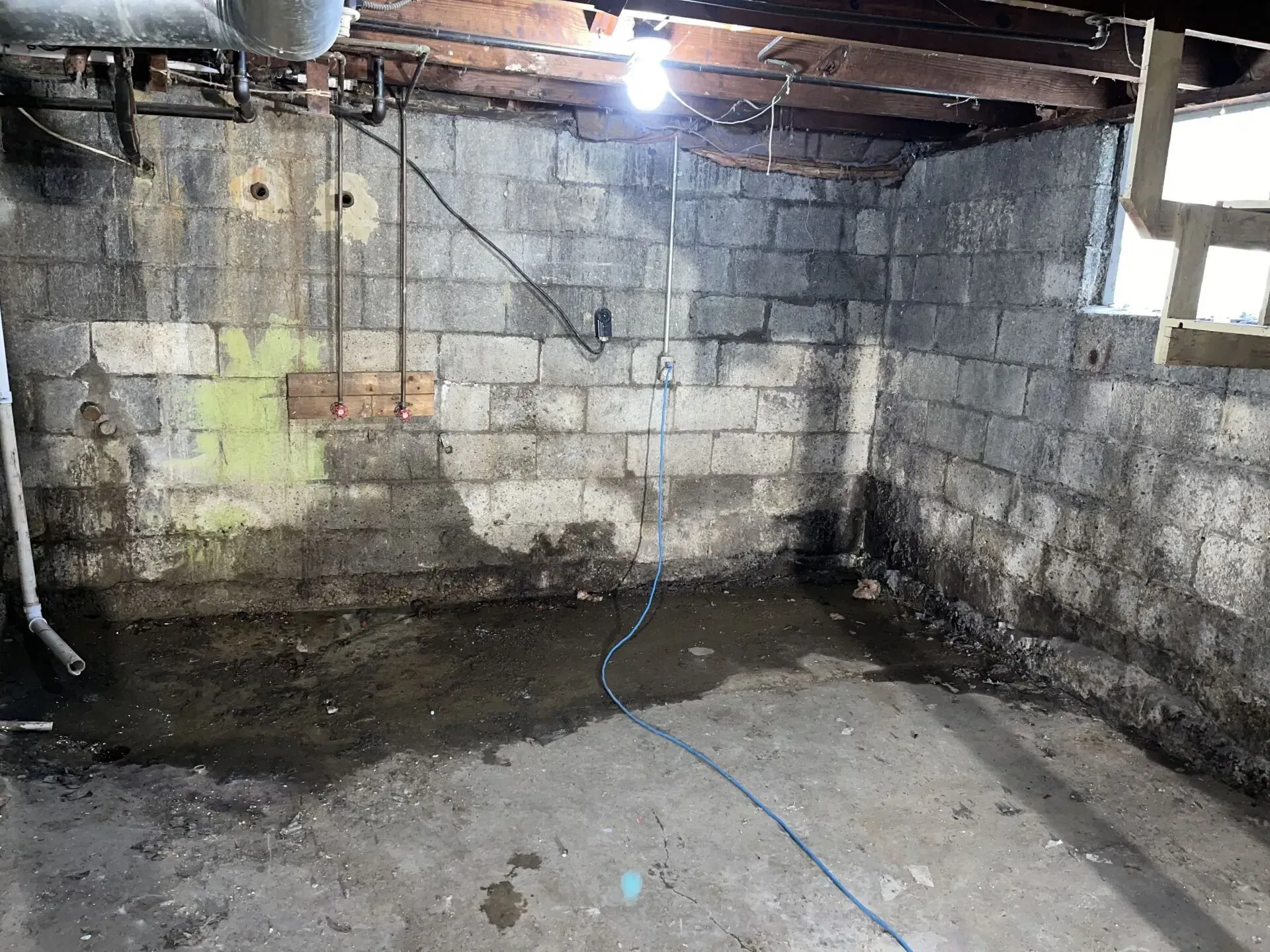Damp unfinished basement with cinderblock walls, exposed beams, wet concrete floor, and a blue cable on the ground