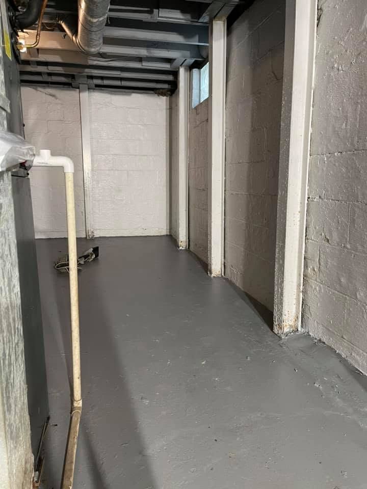 Narrow basement hallway with gray floor, white cinder block walls, exposed ceiling pipes, and a small window