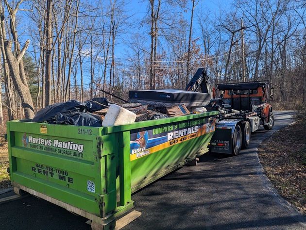 Green dump trailer filled with debris being hauled by a machine on a wooded dirt road
