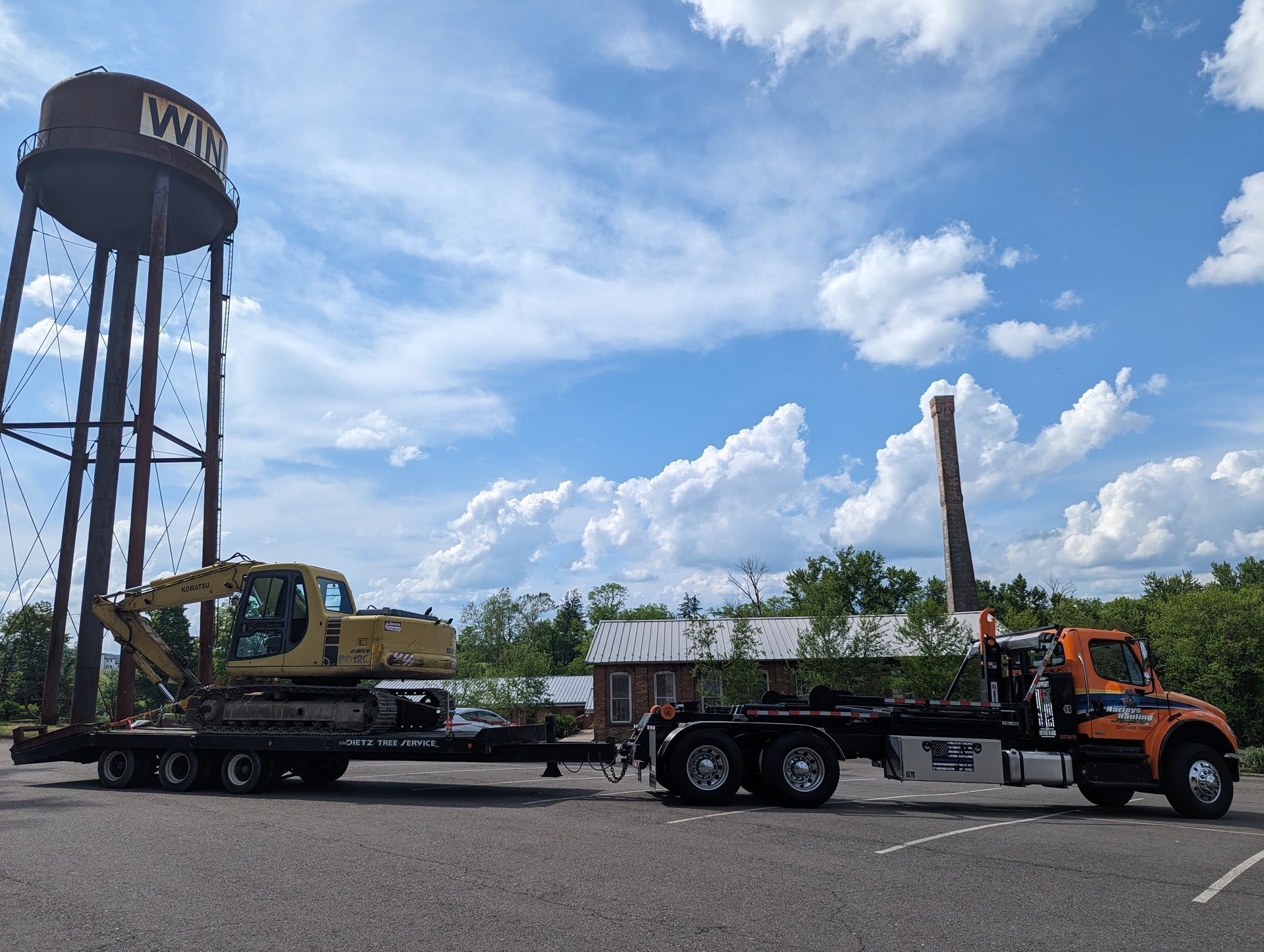 Orange semi hauling construction equipment beside a water tower under a blue sky.