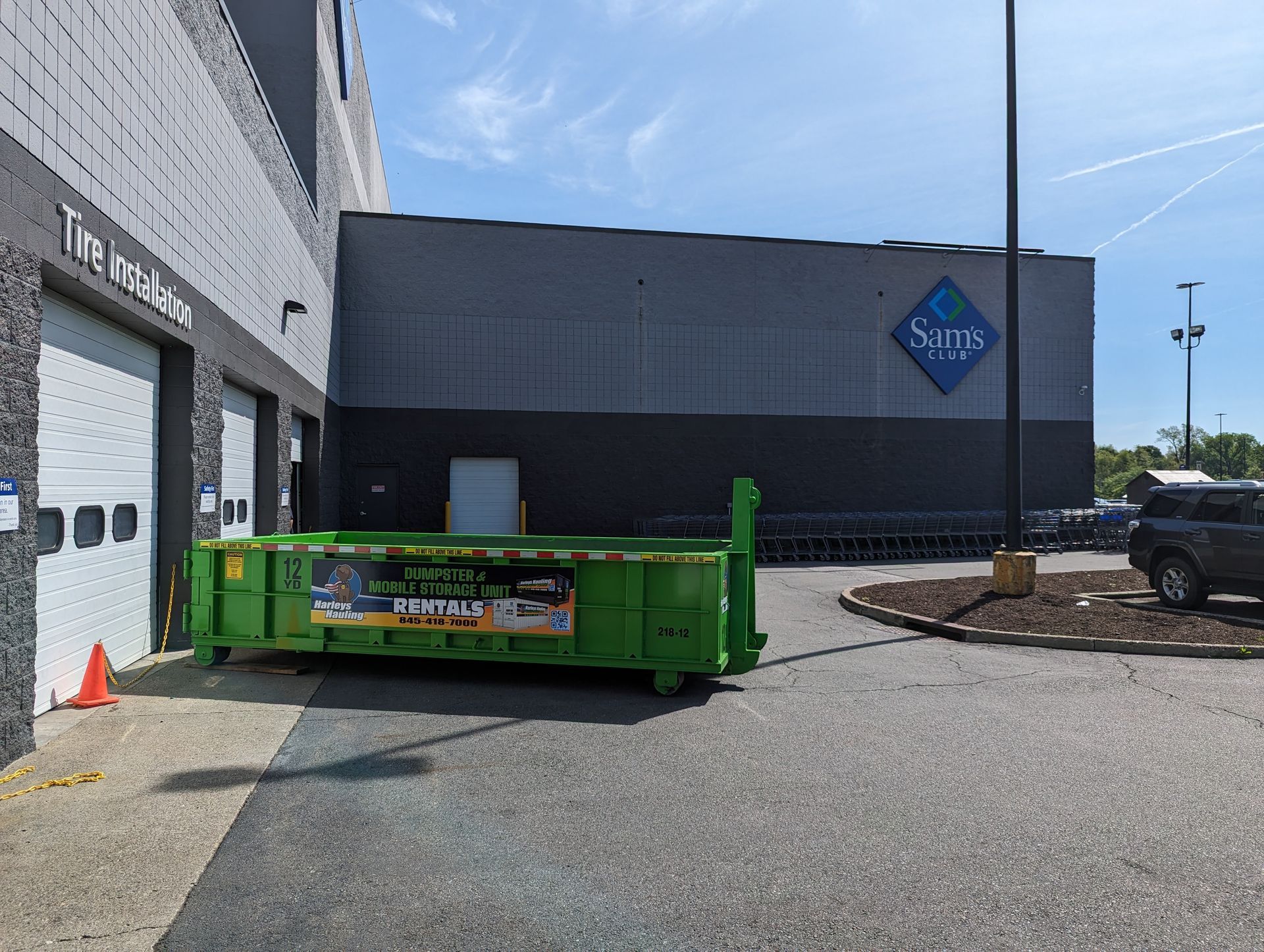 Green dumpster by a warehouse entrance with a blue logo sign and parked truck on a sunny day