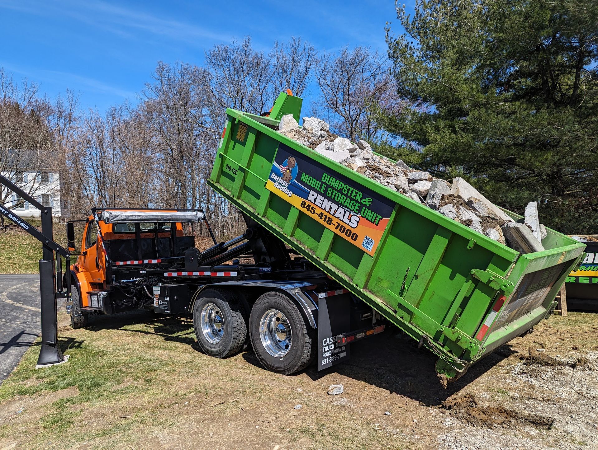 Green dump trailer loaded with rocks, tilted behind an orange construction truck by a roadside
