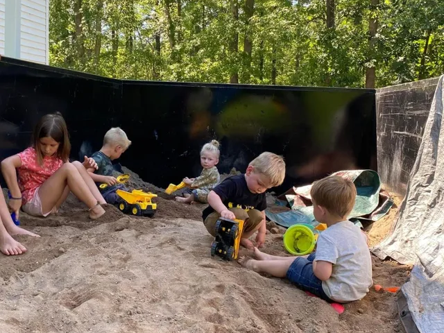 Children playing with toy trucks in a sandbox outdoors, with a black wall and trees behind them