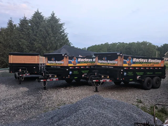 Two black and orange dump trailers parked on gravel in a yard with trees and a house in the background