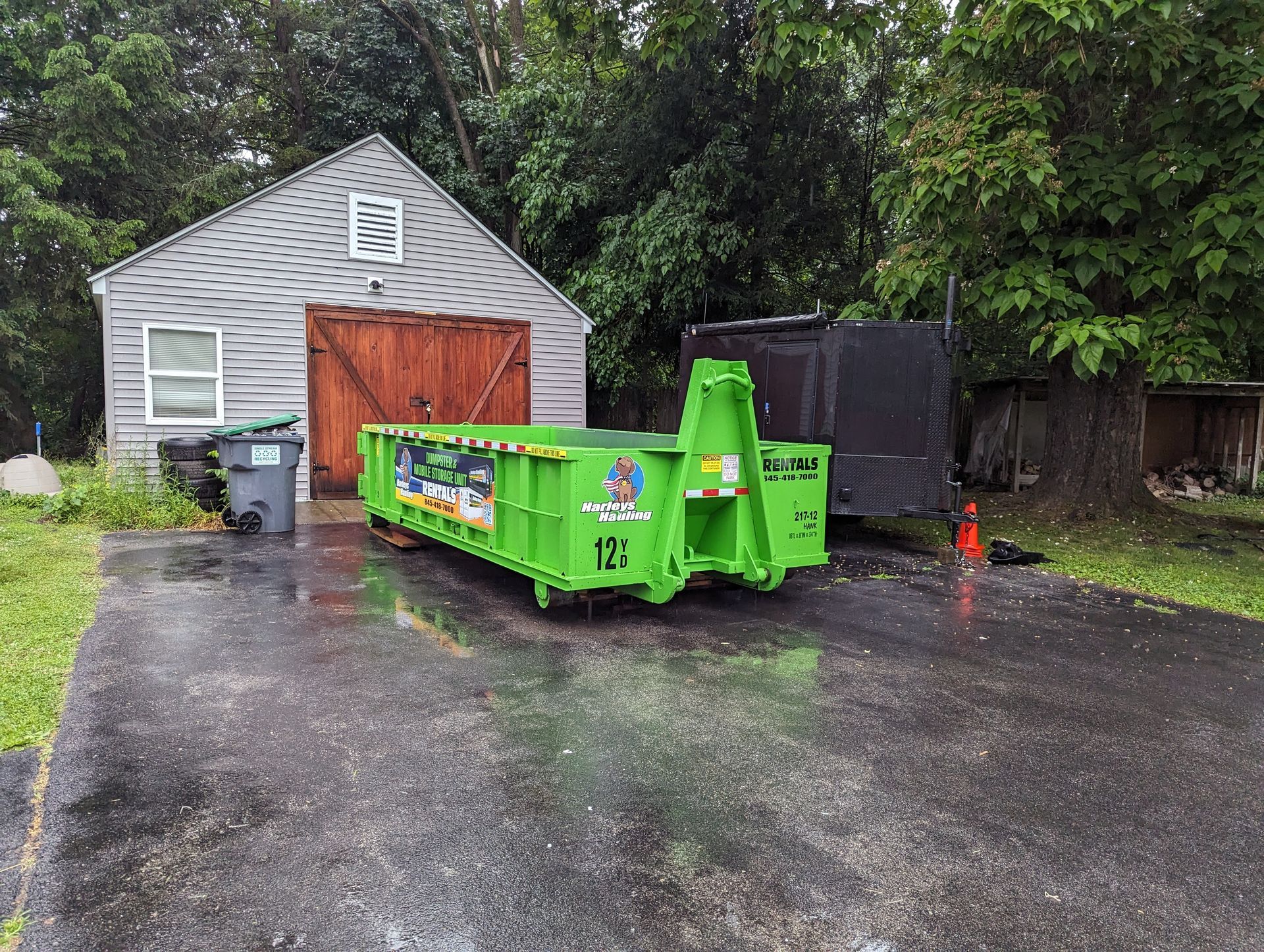 Bright green dumpster on a wet driveway beside a gray shed and trees