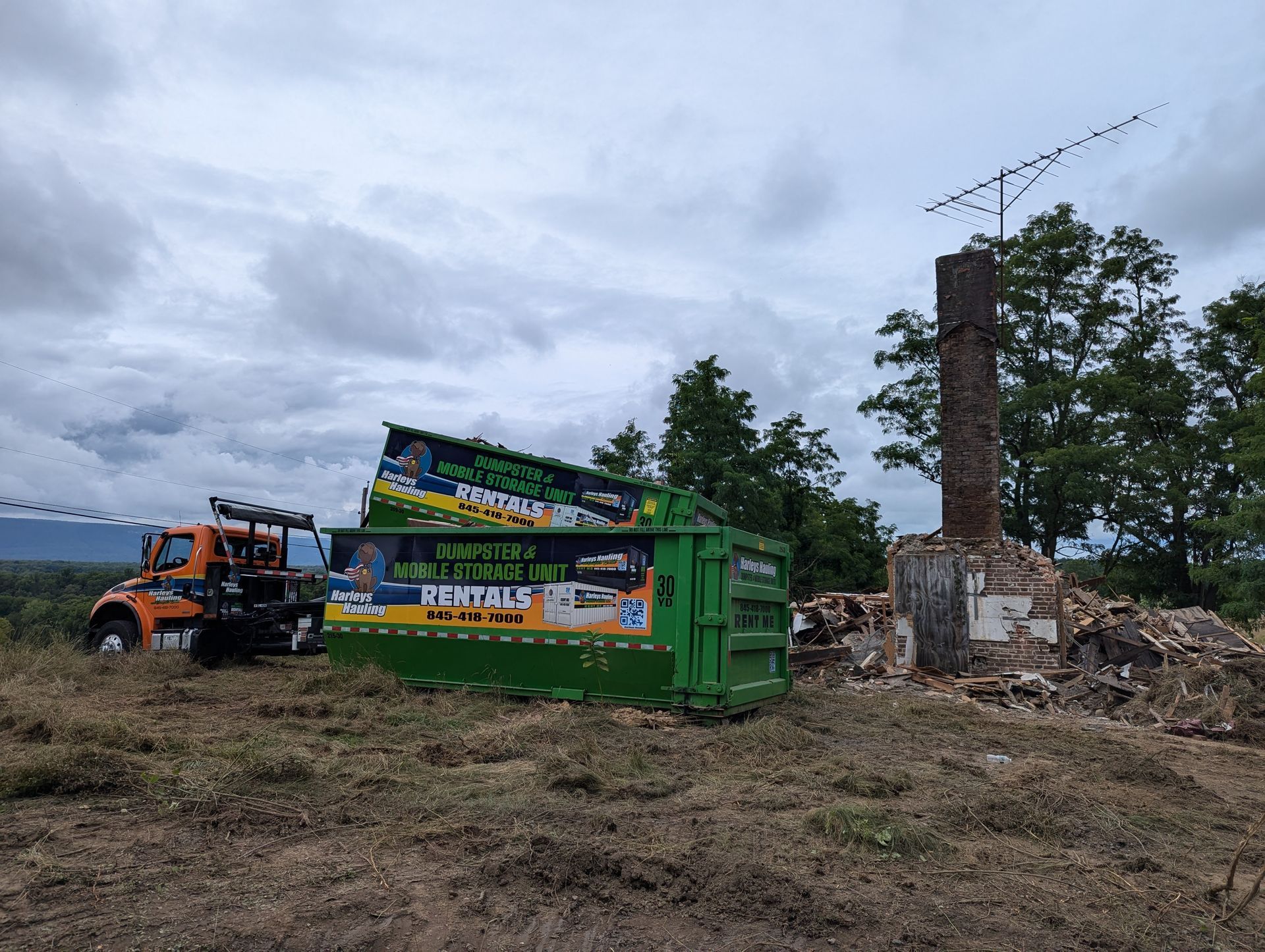 Orange truck and green dumpster at a demolition site with rubble and a tall chimney under cloudy skies