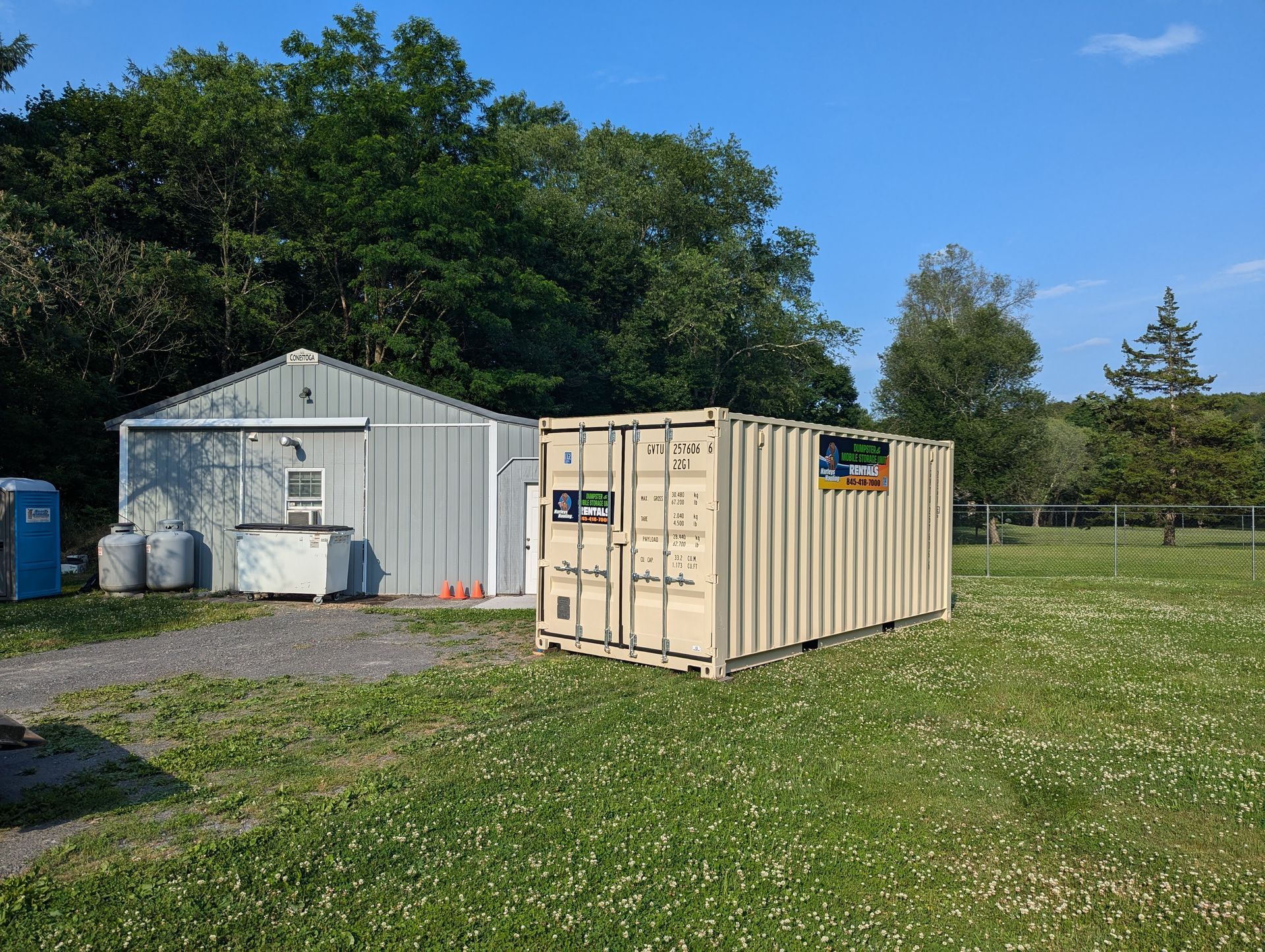 Small metal shed and tan shipping container on a grassy lot under a blue sky