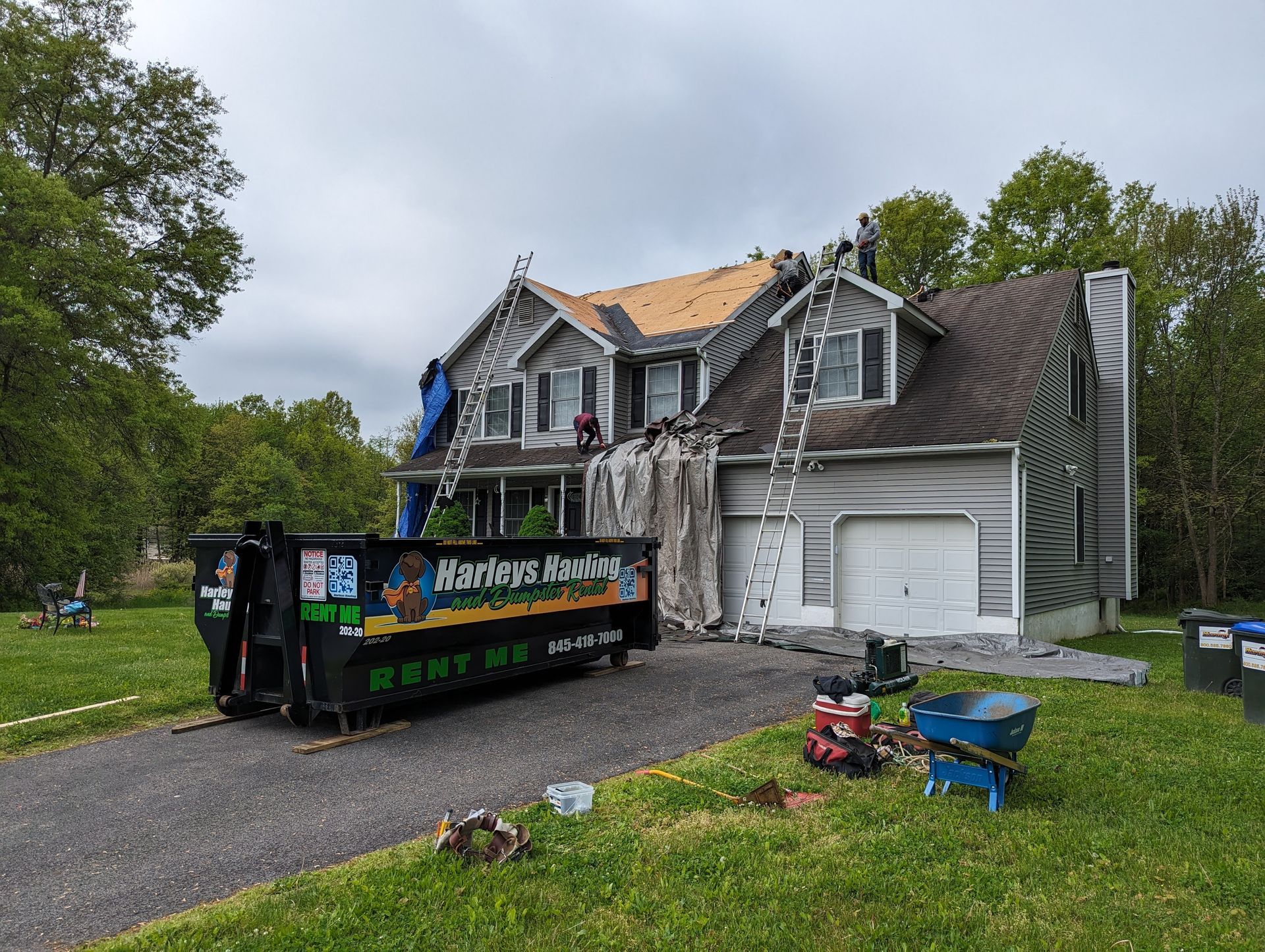 Storm-damaged house with roof repairs, dumpster in driveway, and tarped siding in a suburban yard