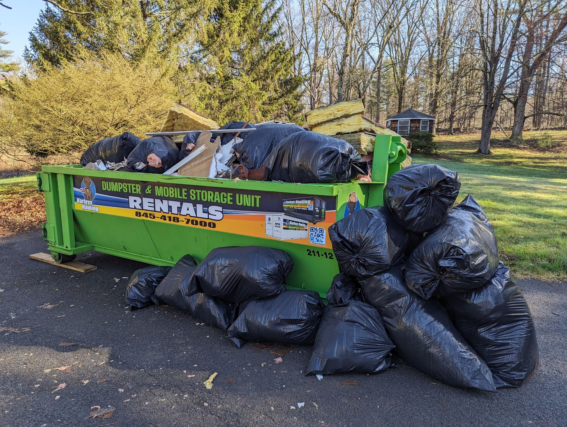 Green rental dumpster filled with black trash bags beside a pile of more bags on a driveway