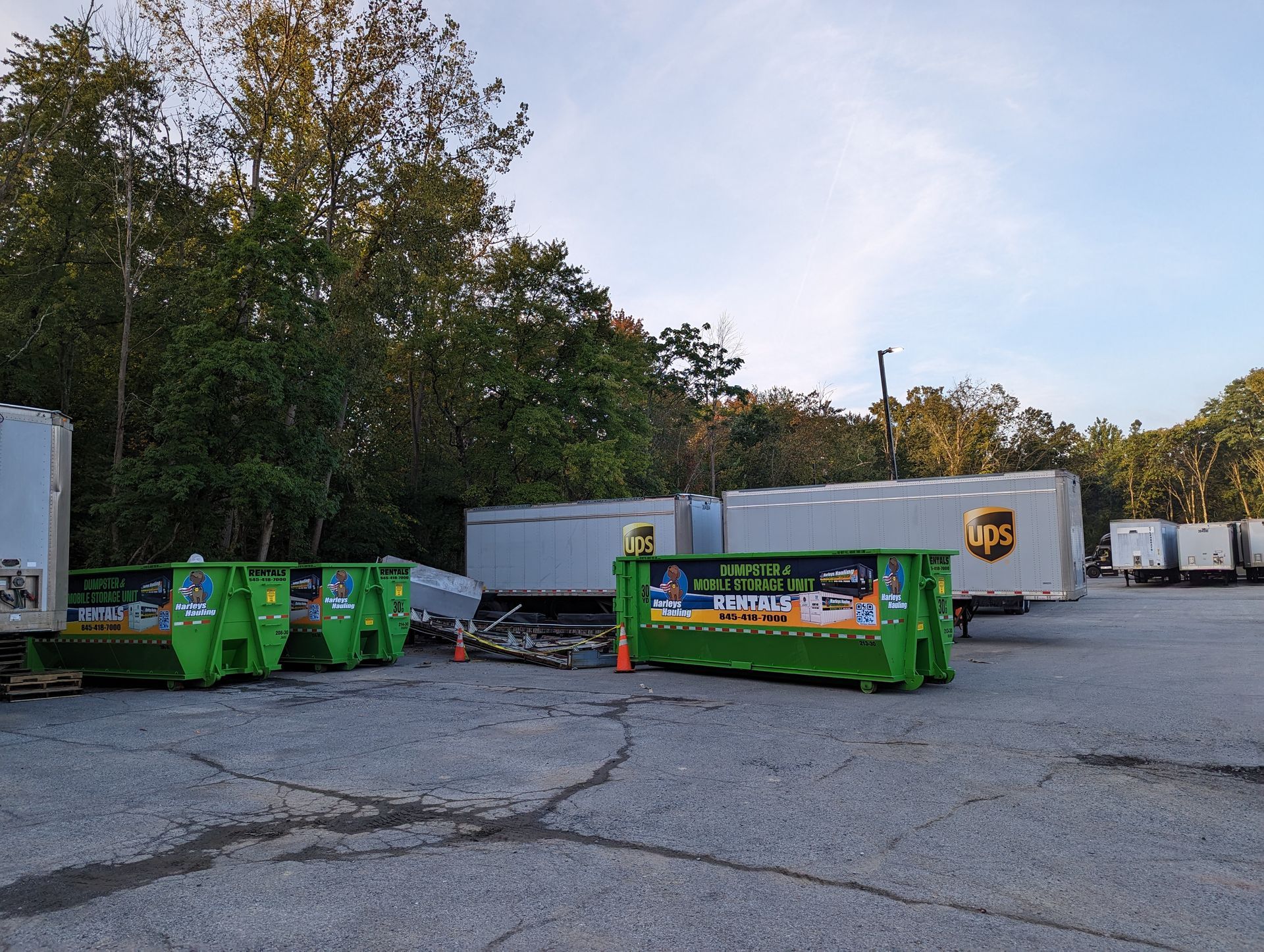 Green dumpsters and white UPS trucks in a parking lot under a cloudy sky.