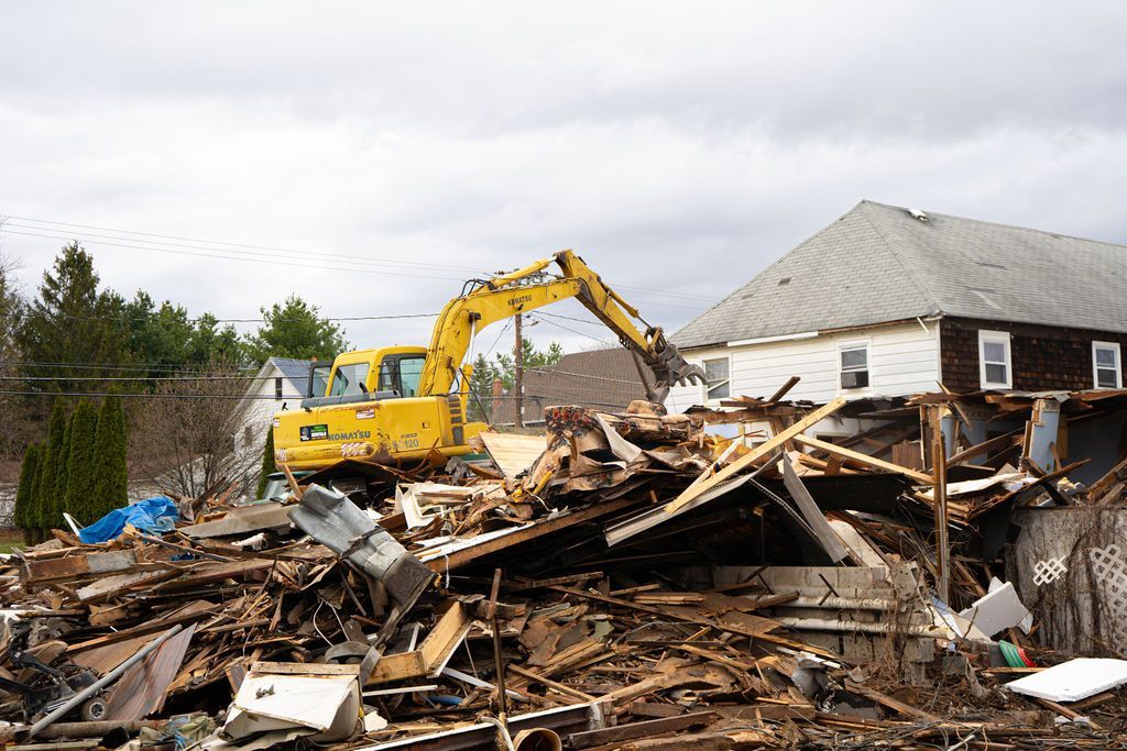 Yellow excavator demolishing a pile of rubble beside a damaged house under a cloudy sky