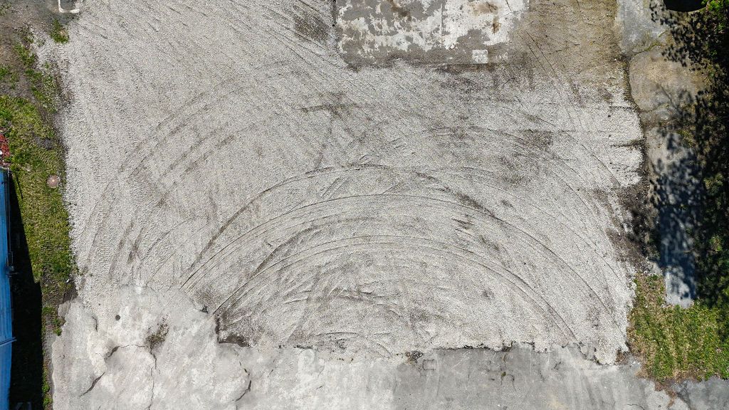Concrete surface with a large circular crack or tire mark, bordered by grass and debris.