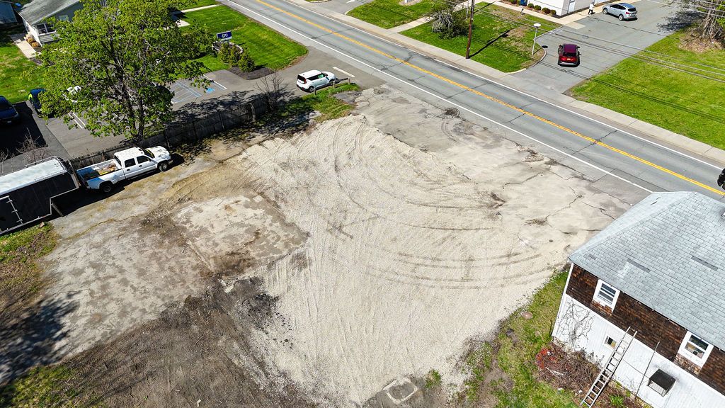 Aerial view of a cleared dirt lot beside a road, with parked vehicles, trees, and a white-roofed building.