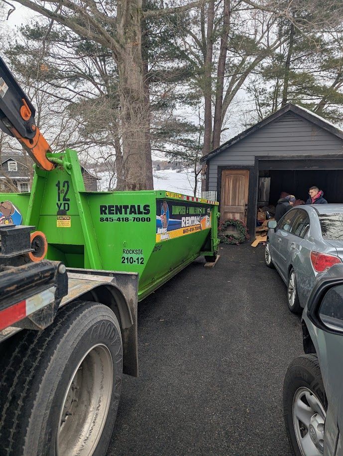 Green rental dumpster truck in a driveway beside a shed, with people unloading debris and a car nearby