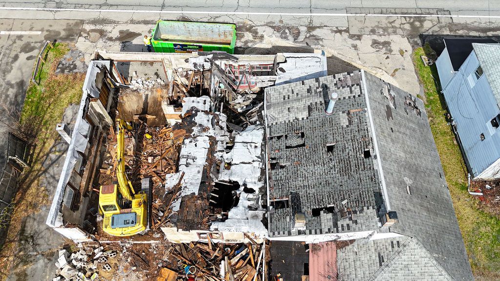 Aerial view of a demolition site with an excavator tearing down a damaged house beside a road.