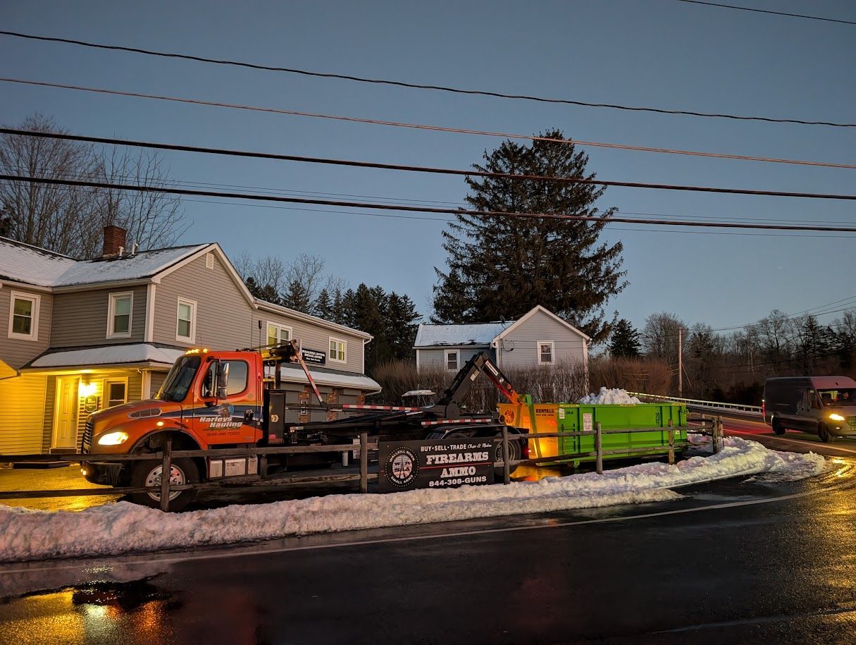 Tow truck with flatbed and green dumpster trailer on a snowy residential street at dusk