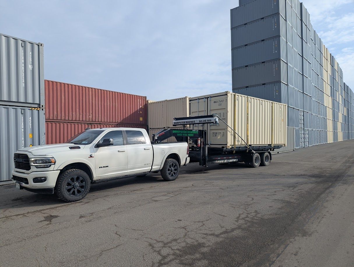 White pickup truck towing a flatbed trailer beside stacked shipping containers in an industrial yard