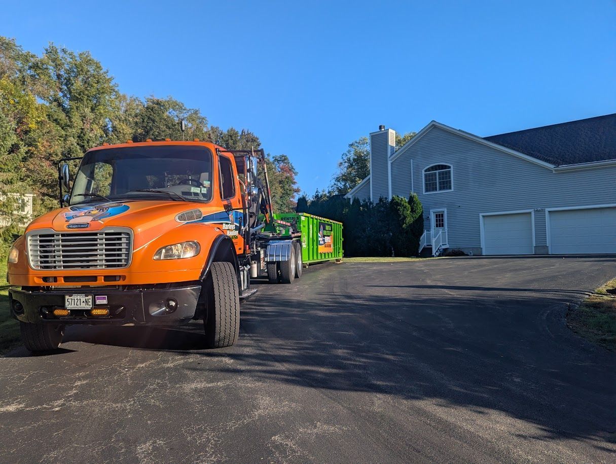 Orange tow truck with green dumpster in front of a white building on a sunny driveway