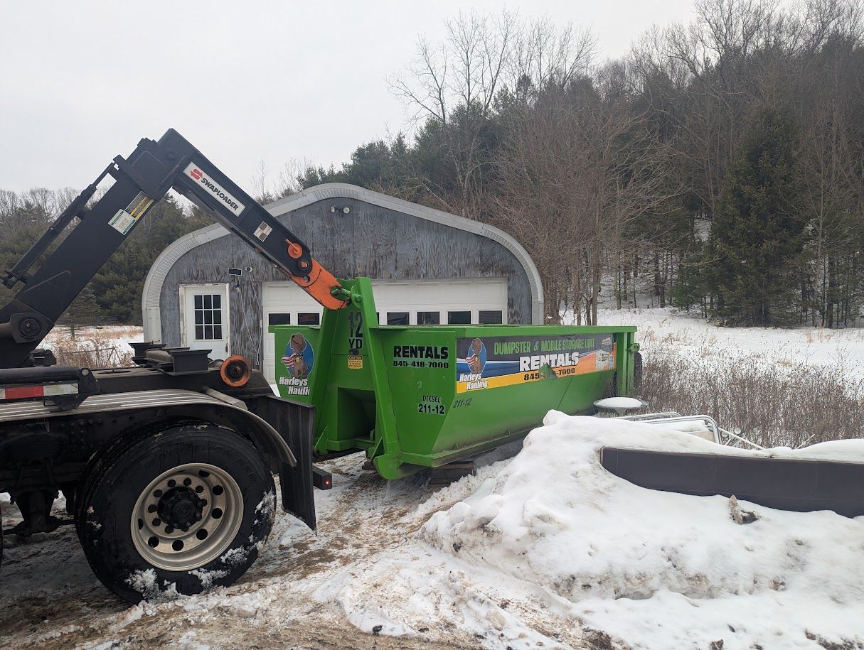 Tractor-mounted snow blower clearing snow beside a green building in a snowy field