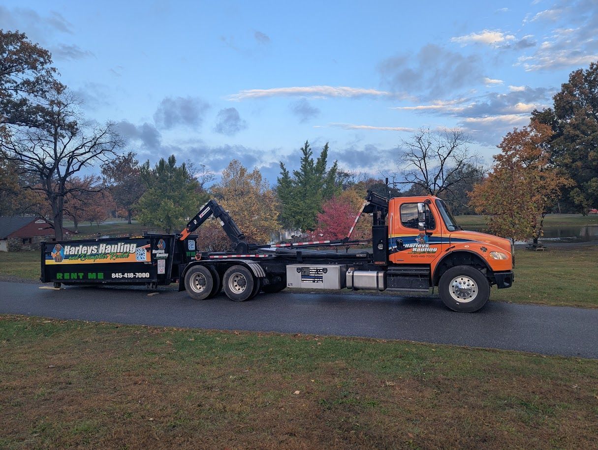 Orange dump truck hauling a green trailer on a road under a cloudy sky in a park