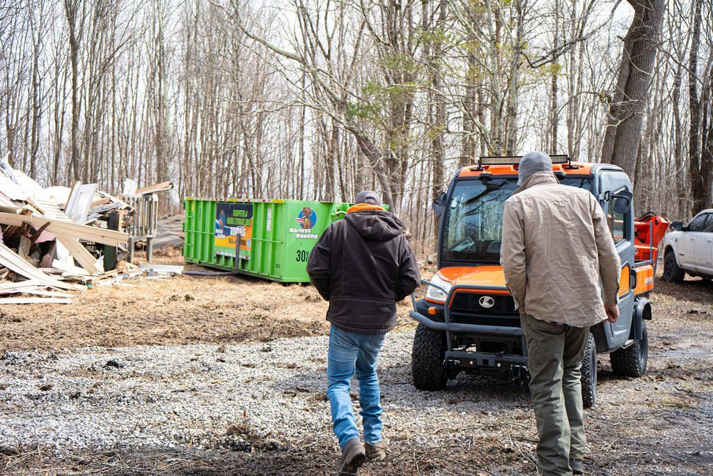 Two people walking toward an orange utility vehicle near a green dumpster in a wooded clearing