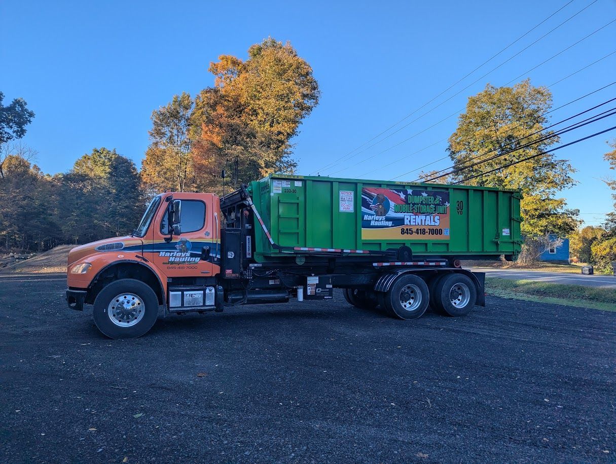Orange truck towing green dumpster trailer on a gravel lot under a blue sky