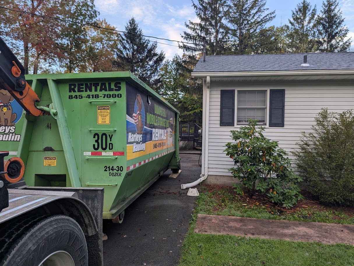 Green dumpster beside a small white house with a worker in an orange jacket and autumn trees in back.